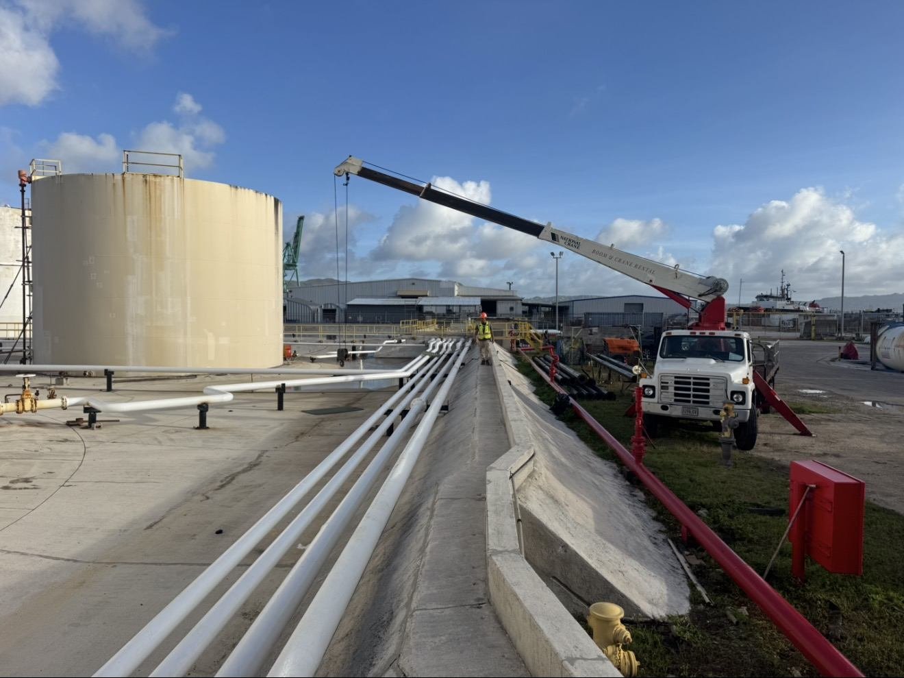 Boom truck lifting beside a fuel storage tank and white pipelines
